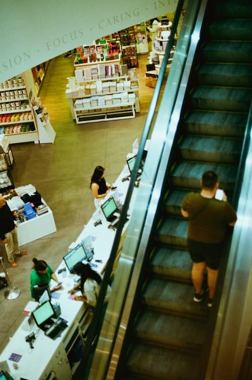 Overhead view of a retail store with a customer riding up an escalator on the right and several people checking out at a counter on the left.